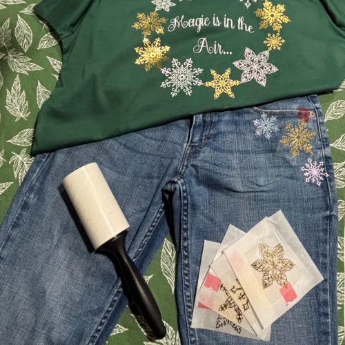 Green shirt with metallic gold, and iridescent white snowflake wreath design, blue jeans, lint roller, and iron-on transfers on a leaf-patterned background.